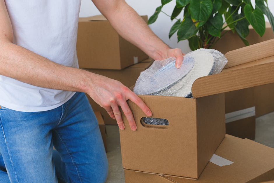 A person wearing a white T-shirt and blue jeans is seen unpacking a fragile item, specifically a glass or ceramic plate, from a cardboard box during a home relocation process. The individual’s hands